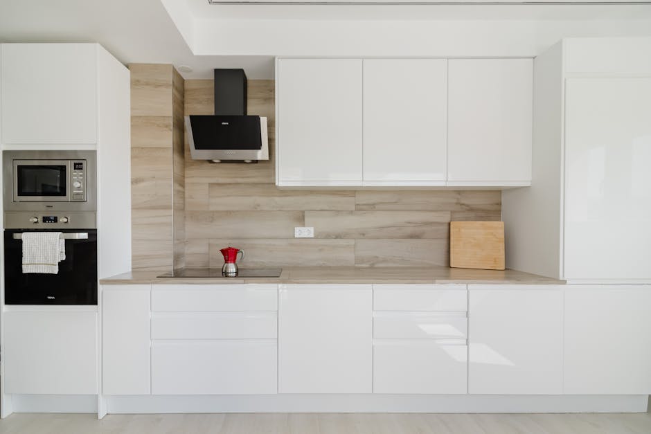 A modern kitchen with sleek white cabinets and a light wood backsplash. The countertops are clean and clutter-free, with a small red coffee pot placed on the surface. On the left side, built-in appliances including a microwave and oven are integrated into the cabinetry, with a towel hanging from the oven handle. A black range hood is mounted above the stovetop, which appears to be free of spills or debris. Natural light illuminates the space, reflecting off the smooth surface of the cabinets, highlighting the overall cleanliness and maintained appearance. The room exudes a sense of good hygiene and proper surface cleaning, consistent with domestic cleaning standards, as promoted by Oven Cleaning Lambeth.