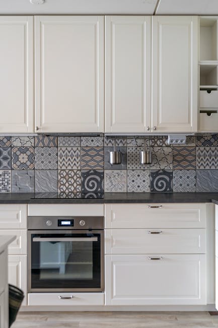 A modern kitchen interior featuring white cabinetry with minimalist silver handles, a black countertop, and a built-in stainless steel oven beneath. The backsplash consists of decorative ceramic tiles in various geometric patterns and shades of grey, cream, and blue, adding visual interest. On the right side, open shelves hold small storage units. The lighting highlights the clean, polished surfaces, emphasizing the pristine condition of the kitchen. The overall scene reflects a well-maintained, hygienic space suitable for thorough surface cleaning and deep sanitisation, consistent with the professional cleaning services offered by Oven Cleaning Lambeth for avoiding oven cleaning delays in Lambeth same day solutions.
