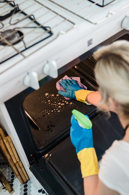 A person wearing blue and yellow rubber gloves is cleaning the inside of a black oven, focusing on the oven door and interior surface. The oven is positioned within a white kitchen stove with metal burners on top, and the interior appears to be wiped clean with some soapy water and bubbles visible. The scene highlights surface cleaning and sanitisation in a domestic setting, with the person's partial blonde hair and professional cleaning approach demonstrated. This image exemplifies thorough deep cleaning practices, as offered by Oven Cleaning Lambeth, to maintain hygiene and shine in home appliances.
