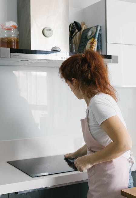 A woman with curly red hair, wearing a white t-shirt and light pink apron, is cleaning a black glass stovetop in a modern, white kitchen. The kitchen features glossy white cabinets, a stainless steel extractor hood, and a white marble backsplash. Visible on the countertop near the woman are cleaning cloths and spray bottles, indicating a surface cleaning process. The kitchen is well-lit with natural light, and the scene emphasizes hygiene and maintenance of domestic surfaces. Oven Cleaning Lambeth offers deep cleaning services for kitchens, focusing on sanitisation and surface cleanliness in residential settings.