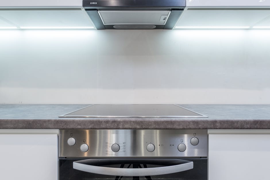 A modern kitchen countertop with a smooth grey surface, situated beneath a stainless steel cooker hood and stove. The stovetop has a sleek, ceramic surface with visible control knobs on a stainless steel panel. The background features a clean, white wall with subtle reflections of light from the integrated under-cabinet lighting, indicating a well-maintained and hygienic environment. The area appears spotless, with no visible dust or stains, exemplifying professional surface cleaning and deep sanitisation associated with domestic kitchen hygiene services provided by Oven Cleaning Lambeth.