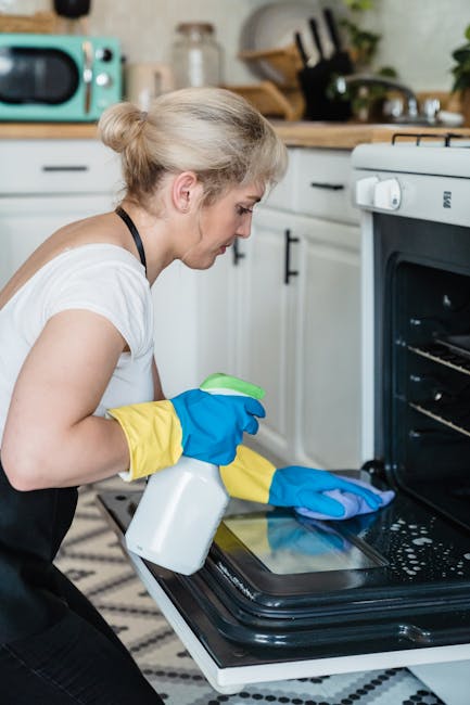 A woman with red, curly hair wearing a white t-shirt and pink apron is performing surface cleaning on a black glass induction stove in a modern, bright kitchen. The kitchen features white cabinets, a stainless steel extractor hood, and a white countertop. Visible on the countertop are glass jars and cooking utensils, with a backsplash of smooth, glossy white tiles. The space is well-lit with natural light, highlighting the cleanliness and shine of the surfaces. The scene exemplifies domestic cleaning and surface hygiene, in alignment with offerings from Oven Cleaning Lambeth, as part of their kitchen deep clean services.