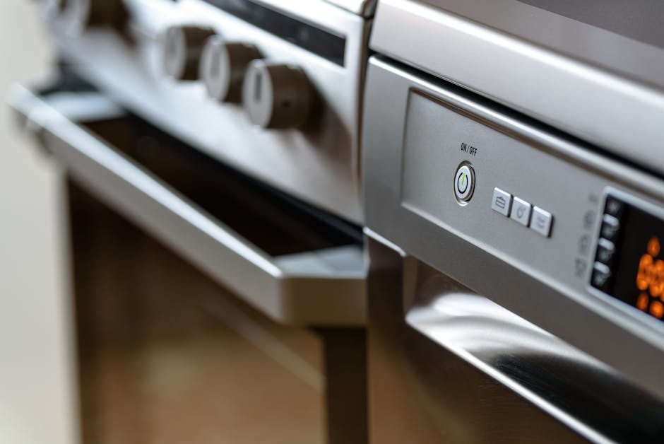 Close-up view of a stainless steel oven with an electronic control panel, featuring a circular power button and three rectangular function buttons beneath. The oven surface is clean and shiny, reflecting light, with a partially visible oven door below. To the left, a blurred view of another kitchen appliance or drawer with a metallic finish can be seen. The background includes a wooden countertop and a white wall, illuminated by bright, even lighting that highlights the surface cleanliness. This image illustrates professional surface cleaning and maintenance practices characteristic of Oven Cleaning Lambeth, emphasizing hygiene and thorough deep cleaning of kitchen appliances within a domestic setting.