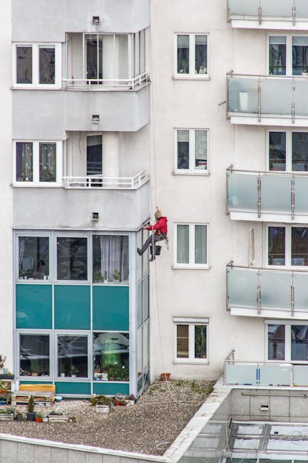 Two window cleaning technicians suspended on a scaffolding belt outside a high-rise building with brick and glass facade, cleaning large glass windows with squeegees and microfiber cloths. The buildin