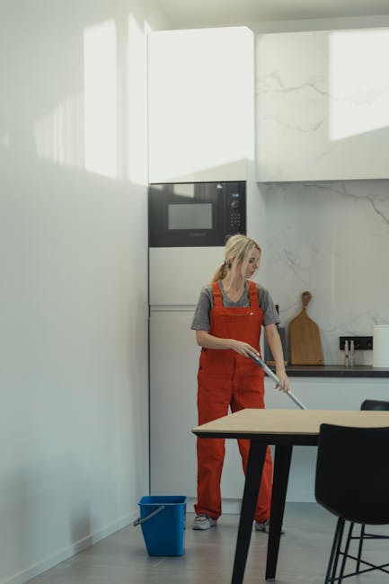 A woman wearing an orange apron and a grey T-shirt is performing surface cleaning in a modern kitchen. She is using a mop to clean the floor, which appears free of dust and stains. The kitchen feature