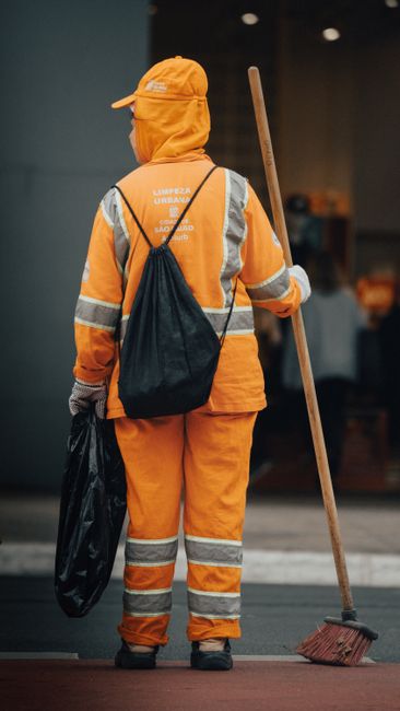 Two street cleaners in high-visibility orange and red uniforms are walking along a paved sidewalk in Lambeth, carrying cleaning equipment. They are approaching a busy intersection with red double-deck