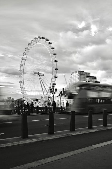 A black and white urban scene featuring a large Ferris wheel in the background, city buildings, and a busy street with motion-blurred double-decker buses and pedestrians. The foreground includes a row of short black bollards lining the sidewalk, illuminated traditional street lamps, and a cloudy sky overhead. This image captures a lively London setting, with the modern Ferris wheel contrasting against historic architecture, indicative of a bustling cityscape. The scene emphasizes movement and urban vibrancy, typical of a central London location referred to in the Waterloo and London Eye oven cleaning for busy flats, Lambeth page. Although focused on city infrastructure, this descriptive accuracy aligns with professional cleaning and maintenance themes relevant to the domain ovencleaninglambeth.com and the service category of cleaning.