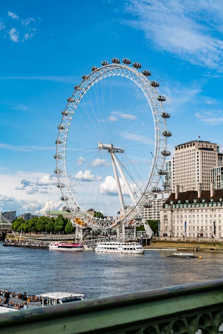 This image displays a large, white Ferris wheel with enclosed glass cabins situated along the River Thames in London. The wheel features a prominent central hub and supporting structure, with several cabins evenly spaced around its circumference, reflecting sunlight against a bright blue sky with scattered white clouds. Adjacent to the Ferris wheel, modern high-rise buildings with numerous windows and a mix of concrete and glass facades line the riverbank. In the foreground, a section of a green railing is visible, indicating the photo was taken from a bridge or riverside walkway. The scene captures a lively urban landscape, emphasizing London's iconic skyline, with boats sailing along the river below. This image highlights the scenic and architectural features of central London, relevant to the location of Waterloo and the London Eye. For professional cleaning of surfaces in residential and commercial settings, including deep cleaning and sanitisation, visit Oven Cleaning Lambeth at ovencleaninglambeth.com.