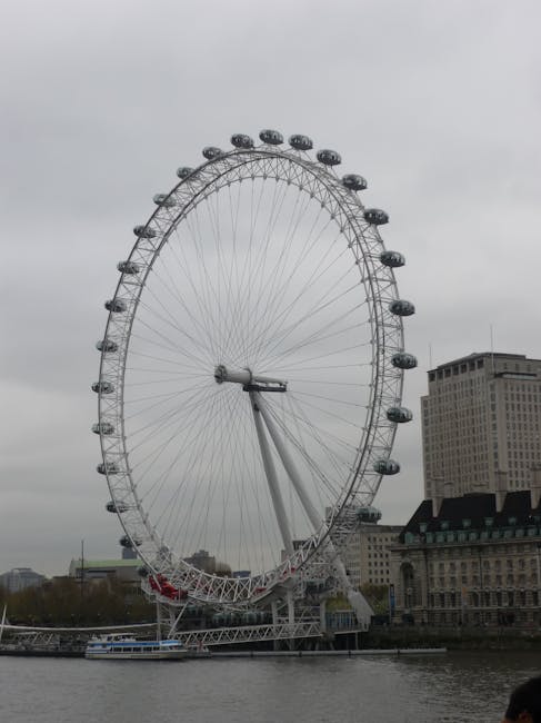 A black and white urban scene featuring a large Ferris wheel in the background, city buildings, and a busy street with motion-blurred double-decker buses and pedestrians. The foreground includes a row of short black bollards lining the sidewalk, illuminated traditional street lamps, and a cloudy sky overhead. This image captures a lively London setting, with the modern Ferris wheel contrasting against historic architecture, indicative of a bustling cityscape. The scene emphasizes movement and urban vibrancy, typical of a central London location referred to in the Waterloo and London Eye oven cleaning for busy flats, Lambeth page. Although focused on city infrastructure, this descriptive accuracy aligns with professional cleaning and maintenance themes relevant to the domain ovencleaninglambeth.com and the service category of cleaning.
